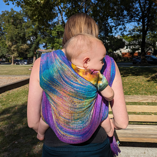 Sarah Labou carrying child in rainbow wrap carrier on her back, Learn Babywearing